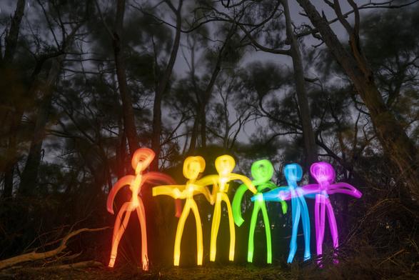 Long-exposure photo taken at night in a forest, featuring six rainbow-colored stick figures made of light, standing hand-in-hand. Each figure glows in a different hue—red, orange, yellow, green, blue, and purple—set against the dark silhouettes of tall, wind-blurred trees under a dusky sky.