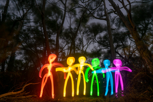 Long-exposure photo taken at night in a forest, featuring six rainbow-colored stick figures made of light, standing hand-in-hand. Each figure glows in a different hue—red, orange, yellow, green, blue, and purple—set against the dark silhouettes of tall, wind-blurred trees under a dusky sky.