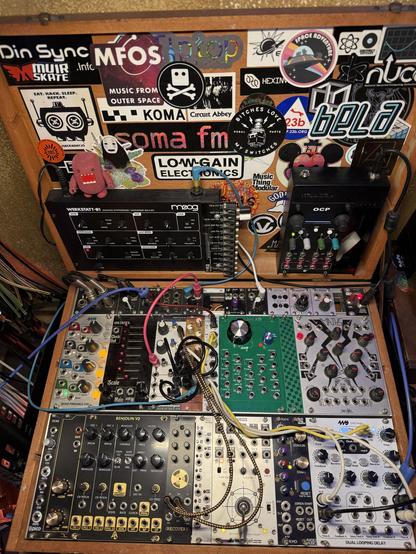 Top down view of a modular case with two rows of full sized synth modules, most of which are not patched. The open lid of the case is plastered in stickers from music companies. Module faceplates range from black and gold to green and silver.
