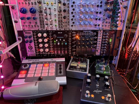 Two full rows of modular synth, unpatched so it is a menagerie of switches and knobs and jacks. The panels on the top row are mostly silver, the panels on the bottom mostly black. On a shelf extending from the bottom are four effects pedals/devices, also unplugged. At the very front is a microphone lying on its velvet red bag.