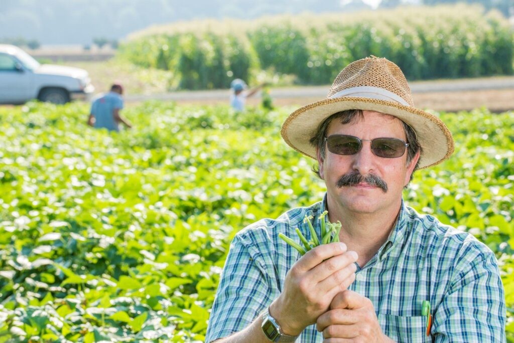 A man wearing a straw hat and sunglasses stands in a lush green field, holding freshly picked beans with more people working in the background.
