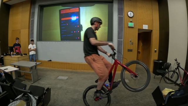 A man is shown performing a wheelie on a red bicycle in a classroom. In the background, a projector is displaying a phone screen running an indistinct app. A man is shown performing a wheelie on a red bicycle in a classroom. In the background, a projector is displaying a phone screen running an indistinct app.