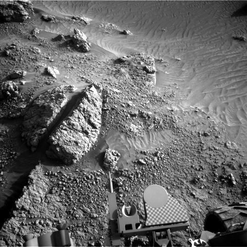 A grayscale photo from the Martian surface shows rough terrain in medium gray, with numerous large and medium-sized rocks emerging from the ground, themselves covered in smaller rocks and gravel. In the upper right corner of the image the ground is much smoother, looking like wind- or water-sculpted sand, and this area appears to be a lower elevation than the rest of the scene. Part of the Curiosity rover is visible in the bottom of the image, including a wheel in the lower-right corner.
