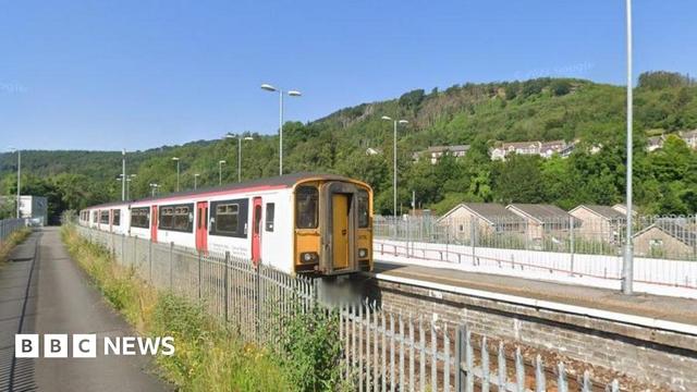 Abercynon train station. A white train with red doors is pulled up at the platform, with a metal fence on the other side of the tracks. A hillside covered in trees can be seen behind it. 