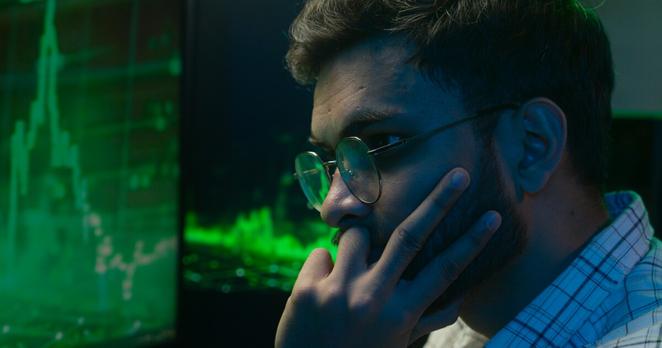 A man wearing glasses looks intently at a screen displaying green financial data and stock charts, suggesting focus on cryptocurrency or digital asset analysis.
