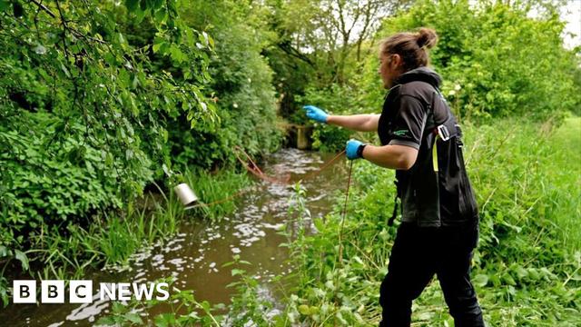 A worker stands on green grass by the banks of a brown stream. He is wearing dark clothes and is swinging a metal cup towards the brown stream on which white frothy bubbles float. There is lush green foliage all around.