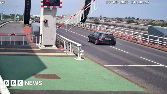 A traffic camera image of a Mini Cooper car passing red safety lights on the Gull Wing Bridge. A barrier is lowering behind the car.