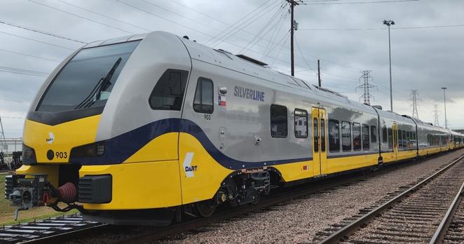 A grey and yellow DART Silver Line train stands on the left track. The right track is empty. The sky behind it is cloudy and there are electrical utility wires visible. A grey and yellow DART Silver Line train stands on the left track. The right track is empty. The sky behind it is cloudy and there are electrical utility wires visible.