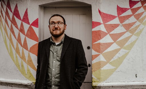 Journalist Mike McGrath-Bryan leans against the toll-booth in St. Luke's, Cork City, which is painted brightly in white and various colours. His photographer has instructed him to look off-camera in a "knowing" or "cheeky" fashion, his eyes pointed skywards.