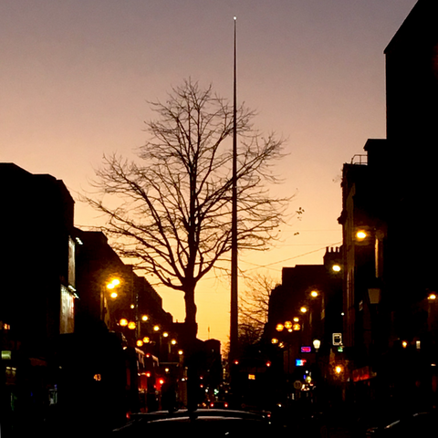 Dusk on Dublin city street. Buildings are dark with street lights and a peach and yellow wintry sunset. A silhouette of a leafless tree is in the foreground and the Spire monument is in the background with a light on top. 