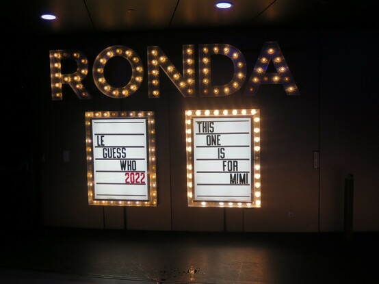 We are looking towards a wall on which in bright lights the word R O N D A is spelled out. Below that are two signs, one saying "Le Guess Who 2022", the other "This one is for Mimi"