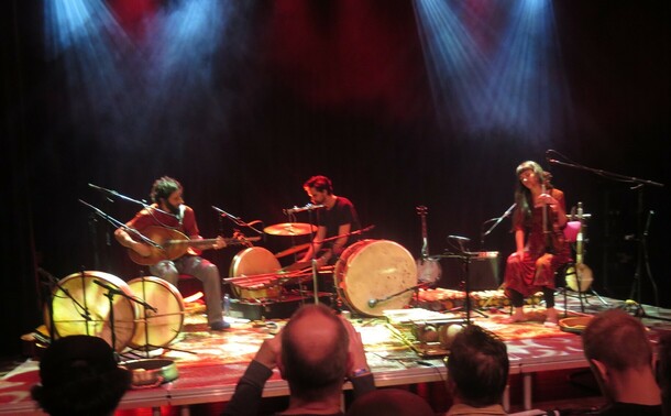 Three musicians on a stage, two men and a woman. The two men are surrounded by drums but only one of them seems to be playing percussion, with the other playing a stringed instrument, possibly a bouzouki. The woman holds a violin but appears not to be playing it. She may be singing. Meanwhile in the foreground a man cursed by male pattern baldness is photographing the musicians.
