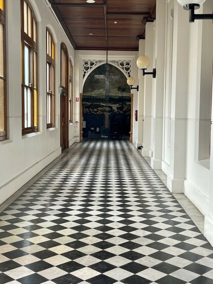 Long formal hallway within a convent, with black and white checkerboard tiles, leading towards a large mural of the Hill of Calvary, with the ghostly shadow of a large cross which has been removed