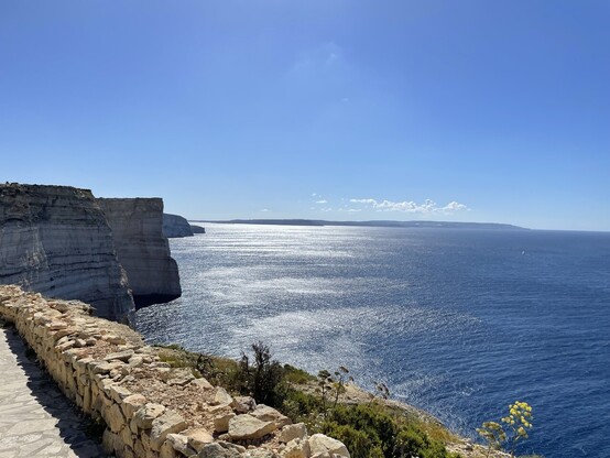 Limestone cliffs on the east coast of Gozo. Blue sky in top half of picture, cliffs to the left, blue sea to the right and the sun shimmering on the water 