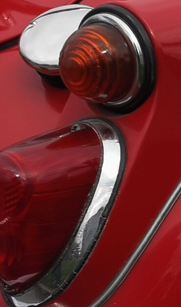 Image shows the rear right hand corner of a red car. It shows a rear brake light with separate indicator, both have Chrome surrounds. Visible just to the left is a bright chrome finished petrol filler cap. 