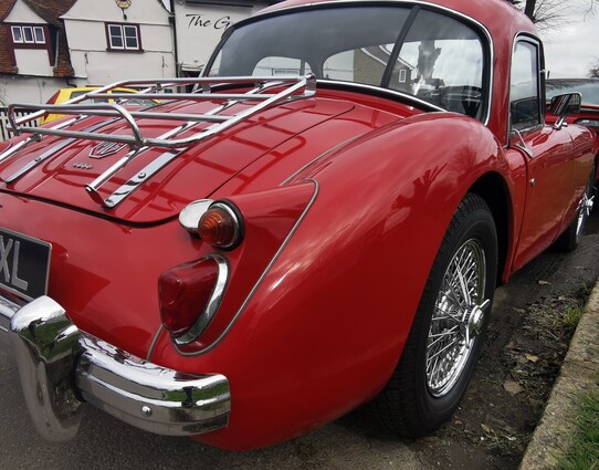 Rear right hand corner of a red MGA. The car has wire wheels, chrome bumper and a luggage rack on the boitlid. It is fitted with a red hard top. 