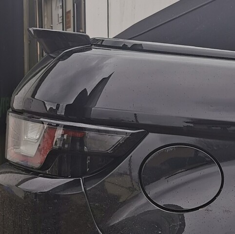Image shows the rear right hand corner of a black car. The car has a black convertible roof and body coloured bumber, there is a lip spoiler at the top of the boot/ base of the roof. The side of the rear light cluster and fuel filler cap are visible. 