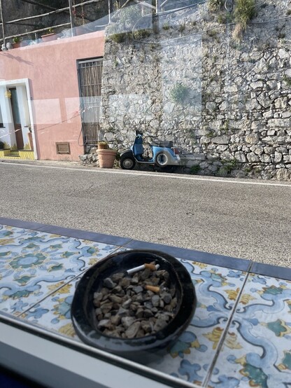 Facade of a grey stone and peach-painted house with blue scooter parked outside; seen through a cafe window with a ceramic tiled window sill outside and an ashtray filled stones with cigarette butts. 