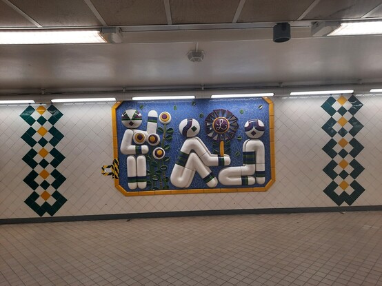 Mural of three robots. One robot waves to the viewer holding what looks like robot sunflowers. One robot sits and gifts another robot a flower in an underground train station in Stockholm, Sweden. 