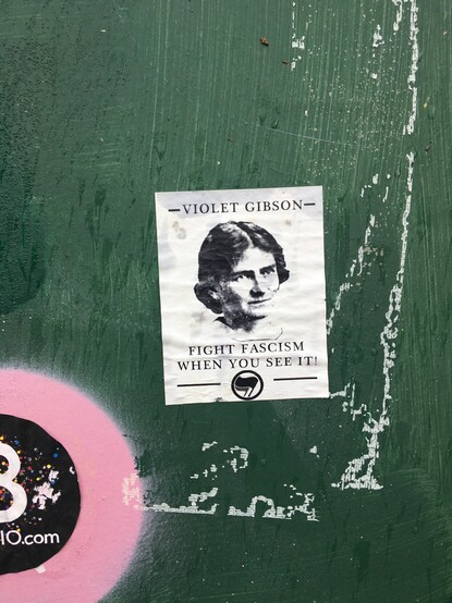 The sticker, on a utility box, showing an illustration of Gibson's face beneath her name and above the phrase "Fight fascism where you see it" along with the double-flag anti-fascism logo. 