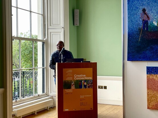 Patrick Mumba at lectern beside large sash window in heritage green walked room with wooden floorboards and his paintings partially in frame. 