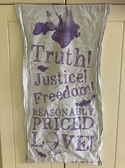 A photograph of a tea towel hanging on a cream cupboard door. The towel is white with lilac drawing and writing. The drawing is a silhouette of a man waving a banner on top of a barricade. Under that is ‘The Glorious 25th May’. In larger writing are the words ‘Truth! Justice! Freedom! Reasonably Priced Love!’ The letter o in the word love is actually a picture of a hard boiled egg.