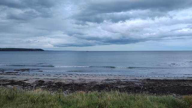 Clonea beach on a nice yet fresh Monday evening. Calm sea at high tide, large white and grey clouds in the sky. Some rain in the back, far out at sea. An old, wooden sailing boat anchored not far away.
Dungarvan, Ireland, July 24, 2023