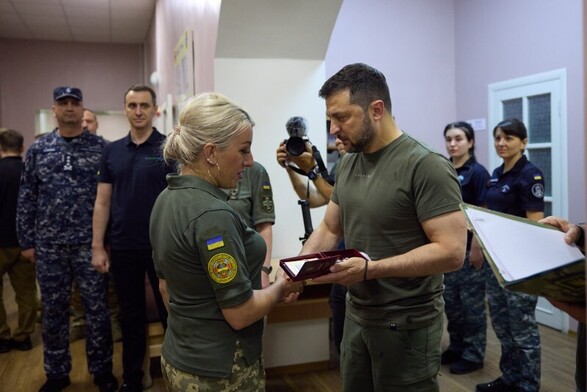 Zelensky handing an award to a female medic whilst shaking her hand. With staff standing in the background. He is looking eyes down with a very serious expression on his face