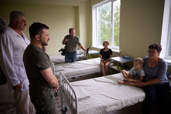 Zelensky standing beside two  hospital bed with a child sitting on a bedwith their mother. Another child is sitting on a second bed with a reporting standing beside her with a microphone .A doctor is also standing beside the president