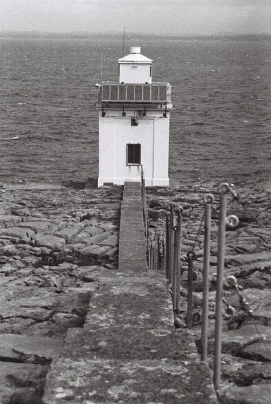 A black and white film photograph of the Black Head lighthouse in County Clare, Ireland. Stone steps descend towards the white lighthouse, a square shaped structure. The seafront is stony, with typical Co. Clare cracked stones - This area used to be a huge river delta, worn down by ice age. Sea is on the background.
