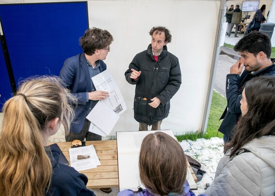 Dr Worthington chatting with members of the public about his research. They are gathered around a bench in a gazebo, with a large book of ancient languages open in front of them. There is also a half-eaten doughnut on the bench. That's from a biomedical engineering demo that was in another tent!