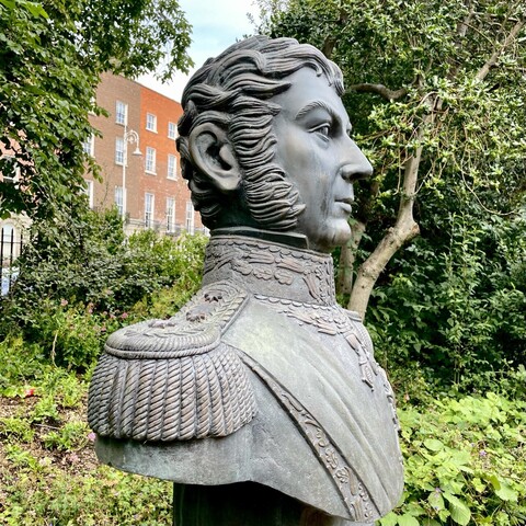 A bronze bust with a matt grey-green colour. A park’s foliage with Georgian-era town houses are visible in the background. The bust is in profile of a man with long sideburns and locks of hair brushed forwards at his head’s temples and crown. He is in a high collared military uniform with shoulder epaulets. The plaque reads: “BERNARDO O'HIGGINS,
BORN CHILE 1776 - DIED PERU 1842. 
LIBERATOR OF CHILE. 
CAPTAIN GENERAL AND STATESMAN. 
THE REPUBLIC OF CHILE TO IRELAND, 1995.”