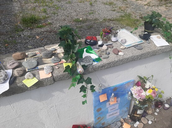 A photo of the front wall on which at places various items including stones with handwritten messages, flowers, post-it notes, artwork, and a copy of the Proclamation of Independence.