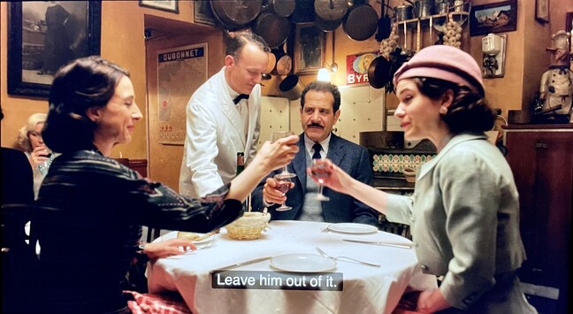 Mrs Maisel’s mother clinks glasses with her daughter, Mrs. Maisel, in a homely Parisian restaurant. On the other side of the round table, Mrs. Maisel’s father is attended by a waiter in a white suit with a black bow tie. He says “Leave him out of it”. 