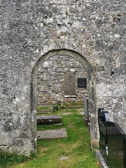 An arched doorway in old church ruins. Above the entrance is a relief sculpture of a sheela-na-gig with a suggestion of facial features that may just be lichen patterns on the stone. Outside the entrance and within the building on the grassy ground are clustered grave slabs and tombstones, some weathered, some relatively new.