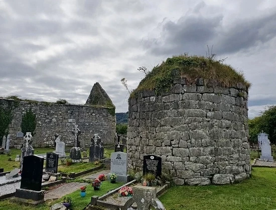 The stump of a round tower in the cemetery outside the old church ruins to the left. The tower is 3 or 4 metres tall and 4 or 5 metres across, rounded on top with grass growing on it. Tombstones surround it, and the sky is moodily overcast.