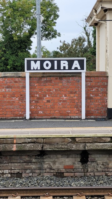 A photo of the name sign at Moira railway station. White capital letters on a black background. The sign has a white border and is held up by two white posts. It sits against a red brick wall. In the foreground is the edge of the platform and the track.