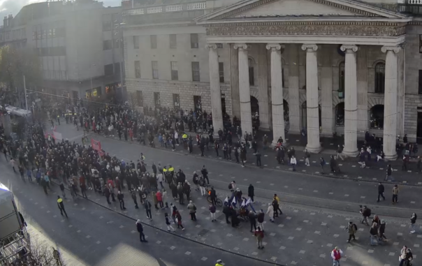 Screenshot from a webcam on O'Connell Street, Dublin, which takes in the GPO and the roadway in front of it. Large crowds are shown gathering along the footpath and also in the wide median which runs along the centre of the street.