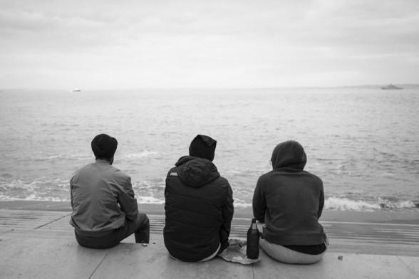 Three people sit on the steps on the bank of a river. A bottle of drink sits between them.