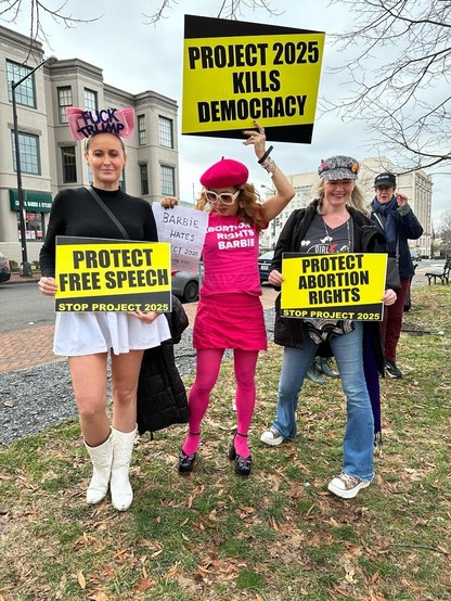 3 women with protest signs. Woman with headress that reads Fuck Trump and a sign that says Protect Free Speech . Woman all in pink with a tee that reads Abortion Rights Barbie holding a sign Pdoject 2025 Kills Democracy and me in a cap covered in buttons holding a sign that reads protect abortion rights.