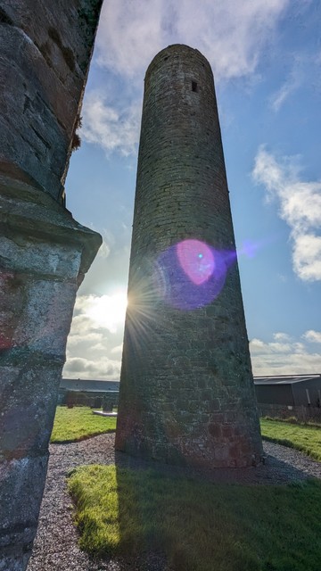 Taghadoe round tower (central) shot looking upward with a church supporting castellated buttress (left) and a glimpse of mid-morning sun peeping round the lower left side of the tower.