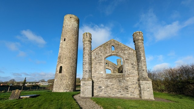 A wide shot of Taghadoe round tower (left) and roofless and windowless church ruin (right) in bright blue-skied and wispy clouded mid-morning sunshine. Rich green grass surrounds the structures and a pebbled path winds up to them.