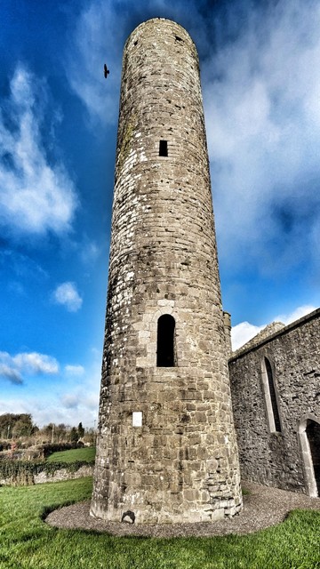 Steep shot from the base of the tower accentuating its main round-headed doorway and mid-height window. The sky has been darkened to rich blue and the coursing of the stonework emphasised. A single black crow flies near, but away from, the top of the tower.