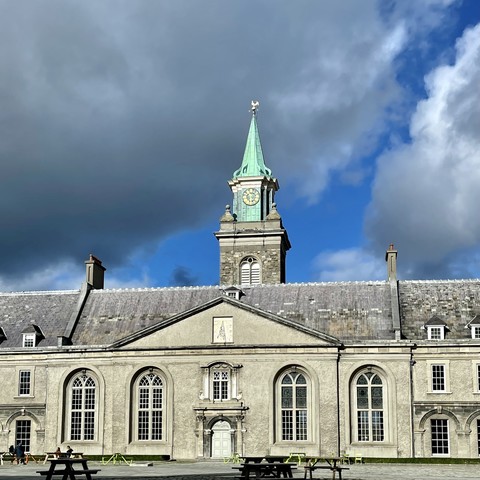 Historic stone building with a clock tower and a spire against a cloudy sky. There are picnic benches in the foreground.