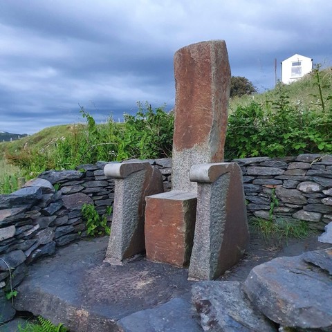 A chair composed of four large blocks of stone. A cubic block which acts as the seat, two sidearms with curved armrests, and a tall back. The chair is carved from red and grey coloured sandstone, and rests in a small alcove set off by a low drystone wall. Vibrant greenery blooms in the background.