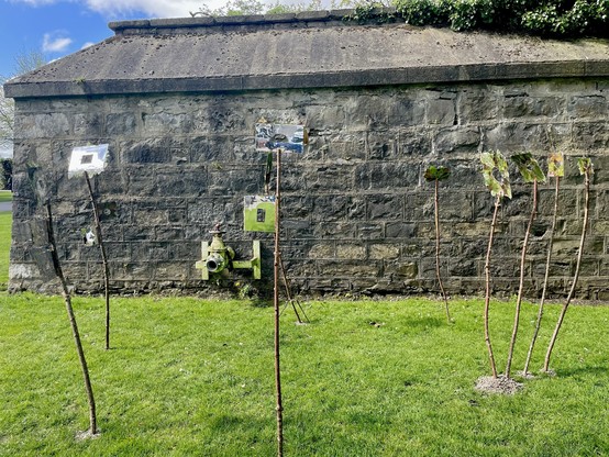 An outdoor art installation with various objects mounted on poles in front of a stone wall, with grass in the foreground.