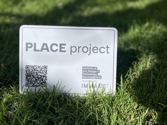 A sign reading "PLACE project" with a QR code lies on grass, indicating an art, design, and technology project by the Institute of Art, Design + Technology Dún Laoghaire in association with IMMA.