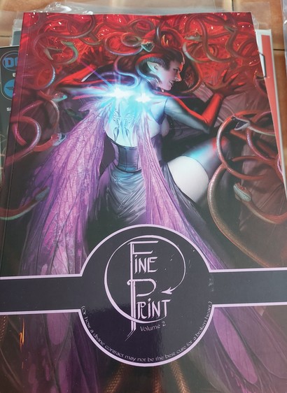 Photo of volume 2 of Fine Print, graphic novel on a stack of other comics on a coffee table. 