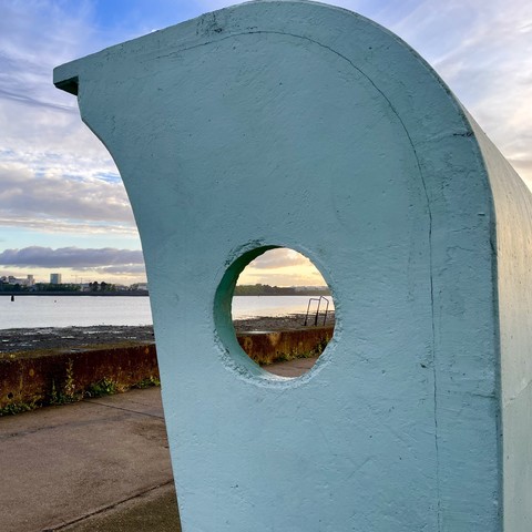 Blue curved structure with a circular hole framing a river and distant buildings at sunset.