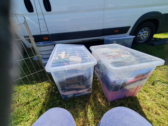 2 knees in blue tracksuit in the centre foreground. Two big plastic tubs full of stuff in front on thr grass. A tub is behind the one on the right. A white van is parked behind that. All on grass. A wire puppy pen is off to the left